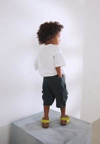 Chemise blanche à manches courtes, short cargo vert foncé avec poches, sandales vertes sur un enfant debout sur un piédestal gris contre un mur blanc.