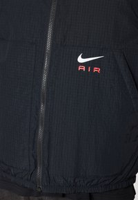 Nike αθλητικό τζάκετ, μαύρο, υφασμάτινο με υφή και ορατό μοτίβο πλέγματος. Διαθέτει φερμουάρ μπροστά, δύο τσέπες και κόκκινο λογότυπο 'AIR'.