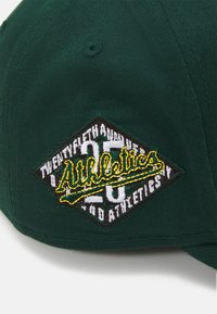 Gorra verde con un logotipo bordado en forma de diamante. El diseño incluye "Athletics" en amarillo con detalles en texto blanco y negro.