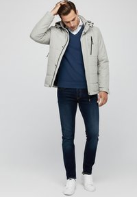 Chaqueta acolchada gris claro con capucha, combinada con un suéter de punto azul marino y jeans oscuros. El modelo lleva zapatillas blancas, mostrando un estilo casual.