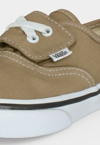 Μπεζ καμβά Vans αθλητικό παπούτσι με λευκά ράμματα, λευκή καουτσούκ σόλα, λευκά κορδόνια και μικρή ετικέτα με το λογότυπο Vans στο πλάι.