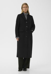 Woman wearing a long black buttoned coat over a gray turtleneck sweater, black pants, and black shoes, standing against a plain white background.