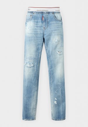 Jeune homme portant un débardeur blanc, un jean clair déchiré, une casquette en jean bleue et des baskets foncées, debout avec une main dans la poche.