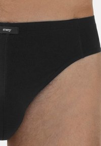 Nahaufnahme einer Person, die schwarze Herrenunterhosen mit einem kleinen Stoffmarkenschild am Bund trägt.