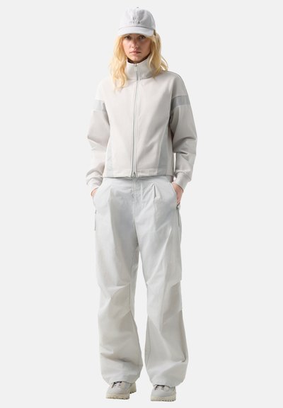 Person wearing light grey zip-up jacket, matching loose pants, white sneakers, and white cap, standing with hands in pockets on white background.