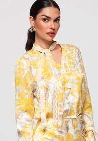 Femme aux cheveux foncés portant un chemisier à fleurs jaune et blanc avec des détails en dentelle et une cravate, portant une grande boucle d'oreille dorée à motif floral.