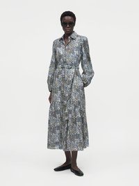 Woman wearing long-sleeve, ankle-length floral dress with a collar, paired with black flats and dark sunglasses, standing against a plain background.