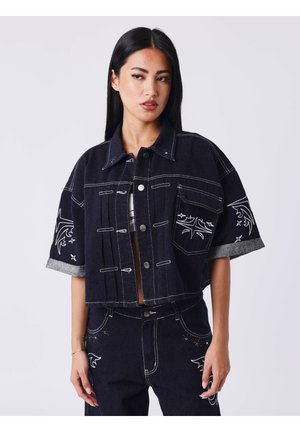 Vrouw die een donker denim jack en broek draagt met witte gestikte ontwerpen en opgerolde mouwen, staand tegen een effen achtergrond.