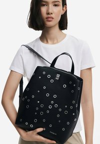 Black leather backpack featuring circular silver eyelets, zip closure, dual handles, and shoulder strap. Brand name "Desigual" visible.