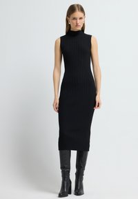 Czarna dzianinowa sukienka z prążkowanej tkaniny, z wysokim kołnierzem i bez rękawów, o dopasowanej sylwetce, kończąca się poniżej kolana.