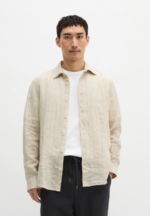 Mężczyzna z krótkimi ciemnymi włosami, ubrany w beżową, pasiastą koszulę na guziki na białej koszulce i czarne spodnie ze sznurkiem, stojący na białym tle.