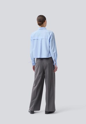 Donna con i capelli raccolti in un bun, che indossa una camicia a maniche lunghe blu chiaro e pantaloni grigi larghi, in piedi di spalle contro uno sfondo neutro.