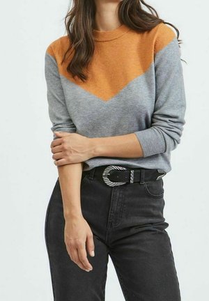 Femme portant un pull à manches longues avec un motif chevron orange et gris, un jean taille haute noir et une ceinture noire avec une boucle argentée.