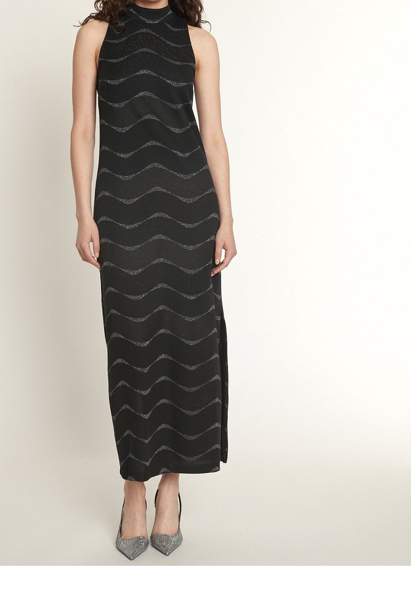 Robe maxi sans manches noire avec un col haut, ornée de motifs métalliques argentés en ondulations et fentes latérales. Associée à des talons hauts brillants.