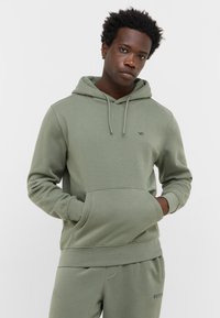 Młody mężczyzna w jasnoszarej bluzie z kapturem i pasujących dresach, stojący z rękami w kieszeniach na jednolitym białym tle.