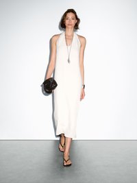 Robe blanche longueur cheville à encolure halter en tissu lisse ; assortie à une pochette tissée noire et des tongs marron. Design simple et élégant.