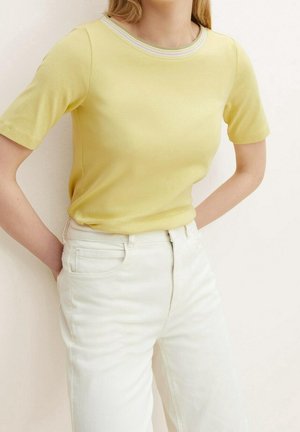 Person trägt ein gelbes Kurzarmhemd mit gestreiftem Ausschnitt, das in hoch taillierte weiße Hosen gesteckt ist, Hände in die Hüften gestützt.