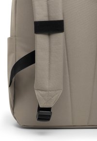 Beige ryggsäck med vadderad baksidan, justerbara svarta remmar, texturerad tyg och triangulär sömnadsdetalj vid remmens bas.