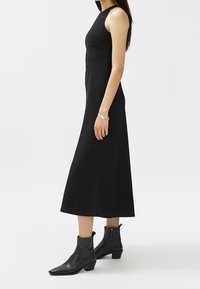 Musta hihaton mekko, jossa on kapea yläosa ja levenevä helma, valmistettu sileästä kankaasta. Yhdistetty mustien nilkkurit, joissa on matala korko.