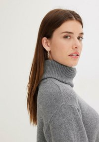 Pull à col roulé gris en maille douce. Il présente un col côtelé et une texture subtile, porté par un modèle aux cheveux longs.