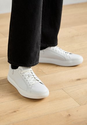Low-top sneakers in white leather, featuring lace-up closure, rounded toe, and minimalistic design. Paired with black, tapered jeans.