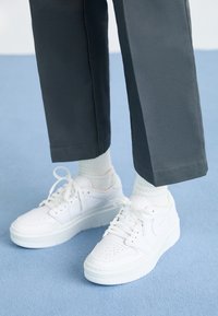 Osoba oblečená v bílých teniskách se šňůrkami, bílých ponožkách a tmavých kalhotách stojí na světle modré kobercové podlaze.