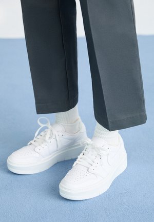 Person klädd i vita sneakers med snören, vita strumpor och mörka byxor stående på ett ljusblått heltäckningsmatta.