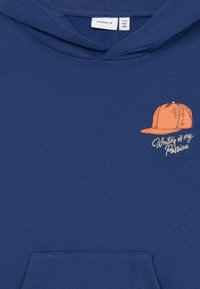 Hoodie bleu avec poche frontale, présentant une illustration de casquette de baseball orange et le texte "Écrire est ma passion" sur la partie poitrine.
