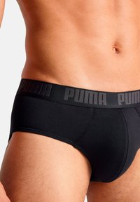 Schwarze Baumwollslips mit einem strukturierten elastischen Bund, auf dem "PUMA" in fettem grauem Schriftzug aufgedruckt ist. Die formschlüssige Passform betont die Körperform.