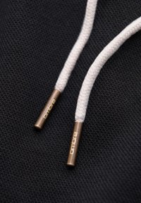 Deux cordons de capuche tissés blancs avec embouts en bronze étiquetés "ombre" sur un tissu noir texturé.
