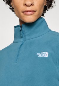 Blue fleece pullover with a zippered neck, featuring a small white logo on the chest. The fabric has a soft texture.