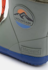 Botas impermeables verdes con suela texturizada azul, que presentan un logo con montañas y un sol. Acentos en naranja brillante en los cordones.