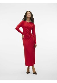 Vestito rosso a maniche lunghe con design drappeggiato, caratterizzato da arricciature su un lato. Tessuto liscio, lungo fino alla caviglia, abbinato a scarpe marroni con punta.