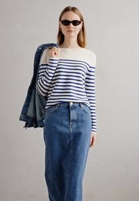 Prugasta plava i bijela bluza dugih rukava i suknja od trapera viskog struka. Model drži traper jaknu. Outfit ima ležeran, opušten kroj.