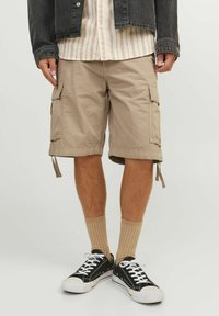 Beige cargo shorts med sidelommer og justerbare snore i kanten, parret med ribbede beige sokker og sorte lave sneakers.