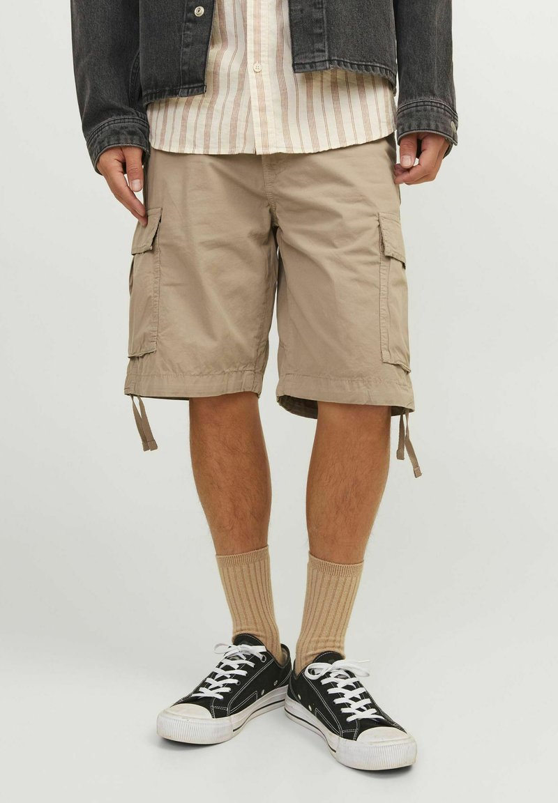 Beige cargo shorts med sidelommer og justerbare snore i kanten, parret med ribbede beige sokker og sorte lave sneakers.