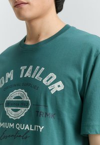 Tirkīzzila kokvilnas T-krekls ar apaļu apkakli, īsām piedurknēm un priekšējo grafisko apdruku ar tekstu un logo baltā un tumši zilā krāsā.