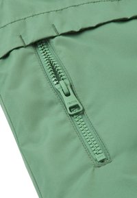 Zelená látka s hladkým povrchom má diagonálny zips. Zipsový ťahák je tiež zelený a označený nápisom "reima".