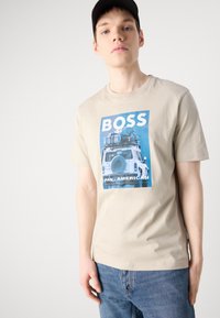 Tricou din bumbac bej cu un imprimeu grafic albastru reprezentând un vehicul cu bagaje, textul "BOSS PAN-AMERICAN" în partea de jos. Croială clasică, guler rotund.