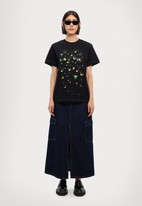 Zwarte katoenen t-shirt met kleurrijke bloemenprint, gecombineerd met een donkere spijkermaxirok met een rits aan de voorkant en grote opgestikte zakken, gedragen met zwarte schoenen.