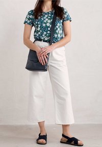 Květinový top s krátkými rukávy v tyrkysové barvě s bílými a zelenými akcenty, spárovaný se širokými béžovými cropped kalhotami a tmavě modrými sandály. Malá crossbody taška.