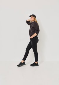Black knit sweater, black fitted jeans, and black sneakers. Model poses with one hand on head, showcasing casual, layered outfit.