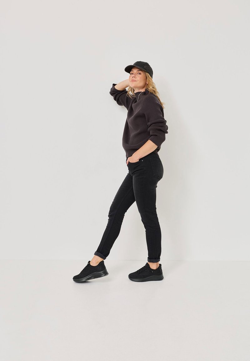 Black knit sweater, black fitted jeans, and black sneakers. Model poses with one hand on head, showcasing casual, layered outfit.