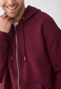 Hoodie zippé bordeaux avec une poche avant, cordons et accent en zip argenté. Tissu doux avec une texture lisse et une coupe décontractée.