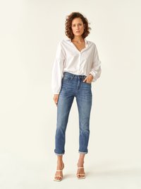 Camisa blanca de botones con mangas voluminosas, combinada con jeans de corte recto en un azul claro y sandalias blancas con tiras. Estilo casual y ajuste relajado.