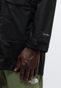 Veste imperméable noire avec une finition texturée. Présente une étiquette "DRYVENT" sur la manche et un poignet avec Velcro. Main reposant sur un pantalon vert.