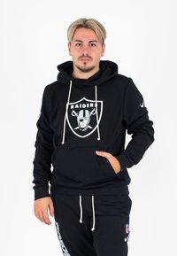 Zwarte hoodie met een Raiders-logo op de voorkant en witte trekkoorden. Voorzien van een kangoeroezak en geribde manchetten, gecombineerd met zwarte broek.
