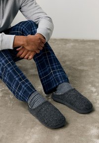 Sivi volneni copati z teksturirano površino in črnim gumijastim podplatom, v kombinaciji s sivo nogavico in karo mornarsko pidžamo hlačami.