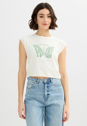 Joven con cabello corto y oscuro, vistiendo un top blanco sin mangas con texto de mariposa y vaqueros azul claro de talle alto.