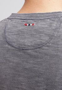 Rückseite eines grauen Strickhemds mit geschwungenen Nähten und einem kleinen horizontalen, rot, weiß und blau gestickten Etikett unter dem Kragen.